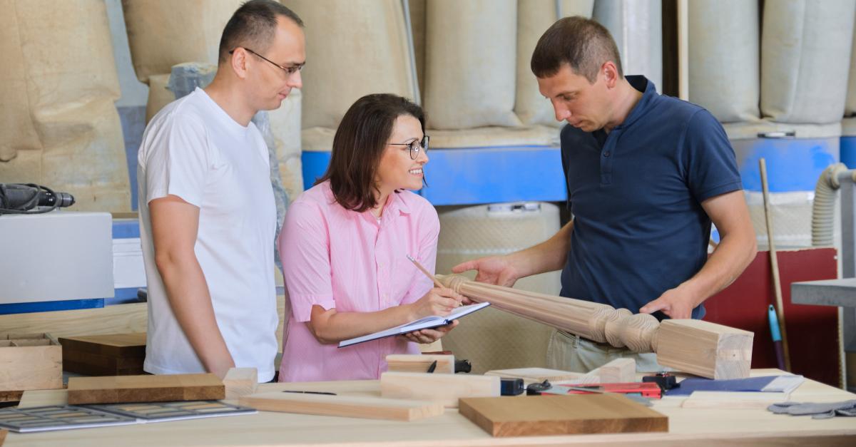 A design professional holding a piece of wood furniture talks with two individuals in a facility.